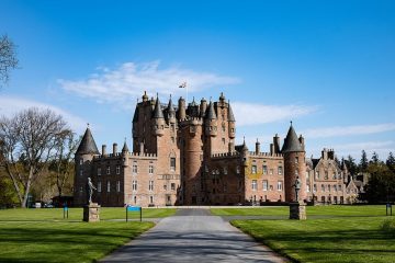 Glamis Castle in Angus, Scotland, a reddish sandstone castle with many turrets and a central tower, seen across a lawn from the approach drive
